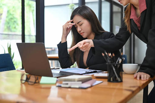 Frustrated businesswoman sitting at a desk with a laptop while a colleague points at the screen, symbolizing financial stress and the importance of money management support like a bookkeeper.