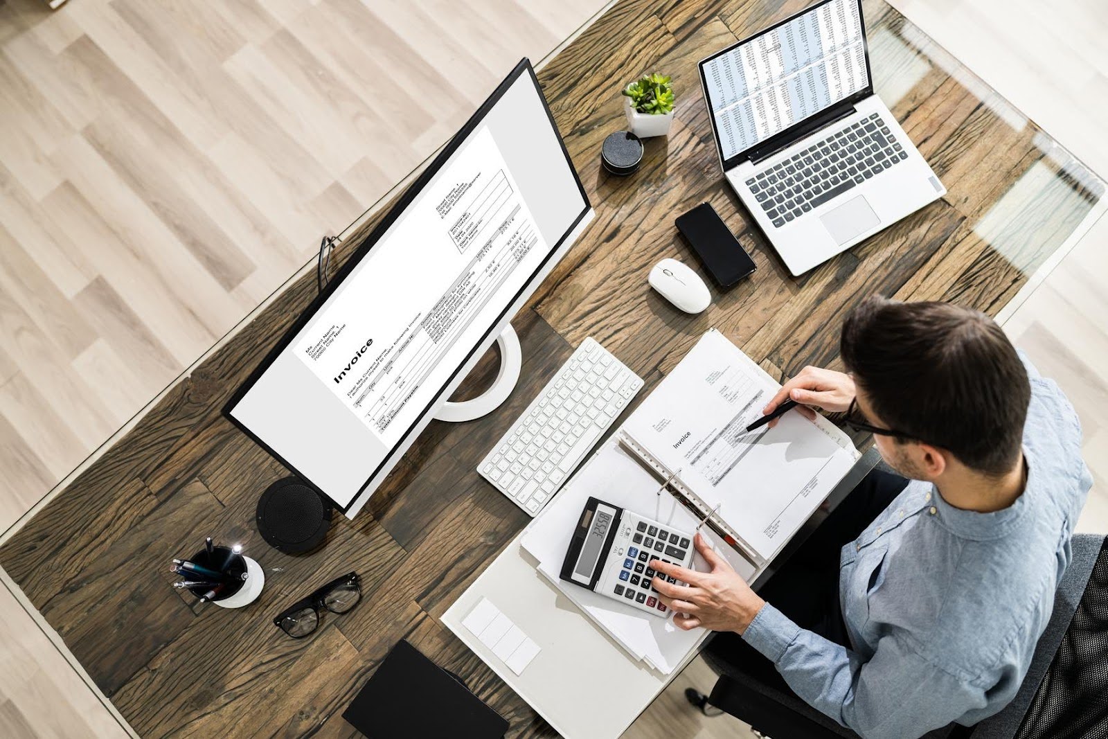 Overhead view of a man reviewing invoices and using a calculator at a modern desk with a desktop monitor and laptop, highlighting common tax-time mistakes small business owners make.