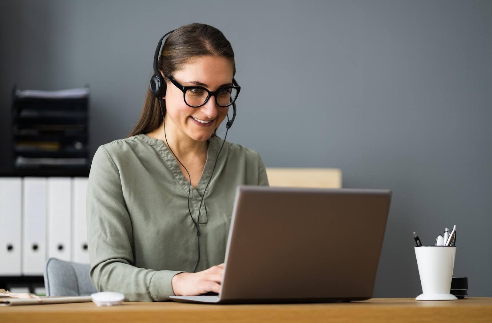 Woman wearing a headset and smiling while working on a laptop, representing the balance of time vs. money when deciding to outsource a role.