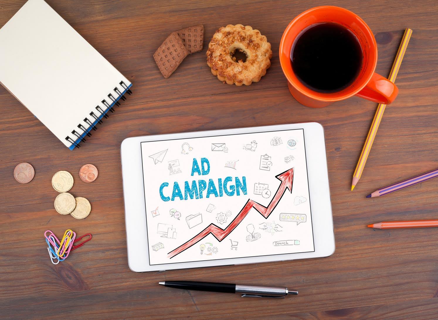 Tablet screen showing ‘Ad Campaign’ with an upward arrow graphic, surrounded by coins, coffee, notebook, and stationery—representing online advertising tools like Facebook Ads and Google Ads for local businesses.