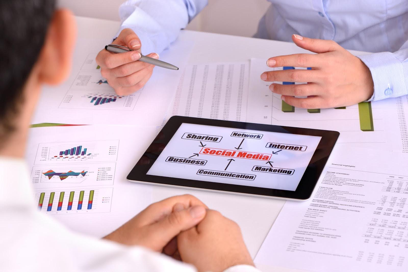 wo professionals reviewing charts and graphs with a tablet displaying a social media concept map, highlighting key metrics such as sharing, network, internet, business, marketing, and communication.
