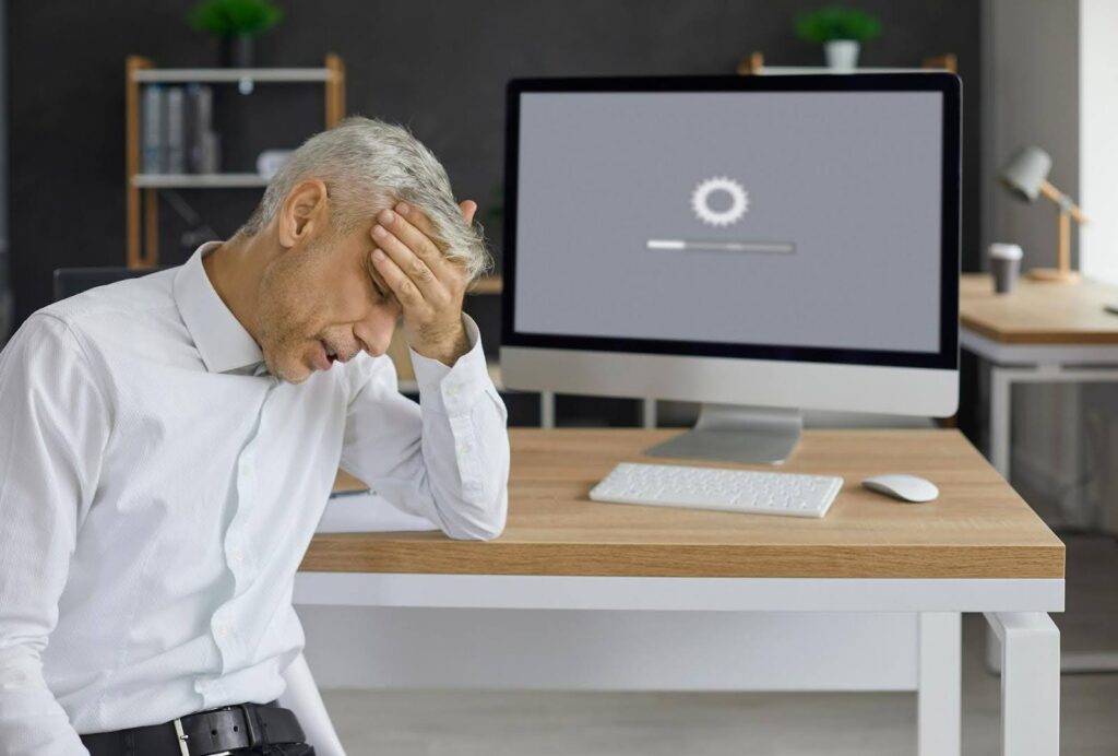 Frustrated businessman sitting at desk with hand on forehead, facing a desktop computer stuck on a loading screen—highlighting the importance of website speed and its impact on user experience and business performance.