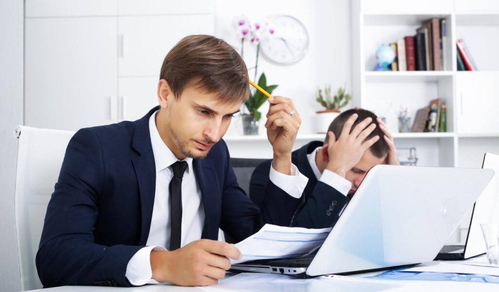 Two stressed businessmen reviewing financial documents on laptops, showing the impact of a bookkeeping mistake on small business finances.