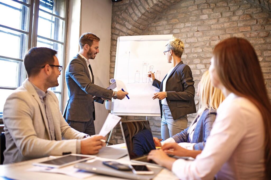 Business team in a meeting with two professionals presenting growth charts on a flip board, representing strategies and challenges of business scaling.