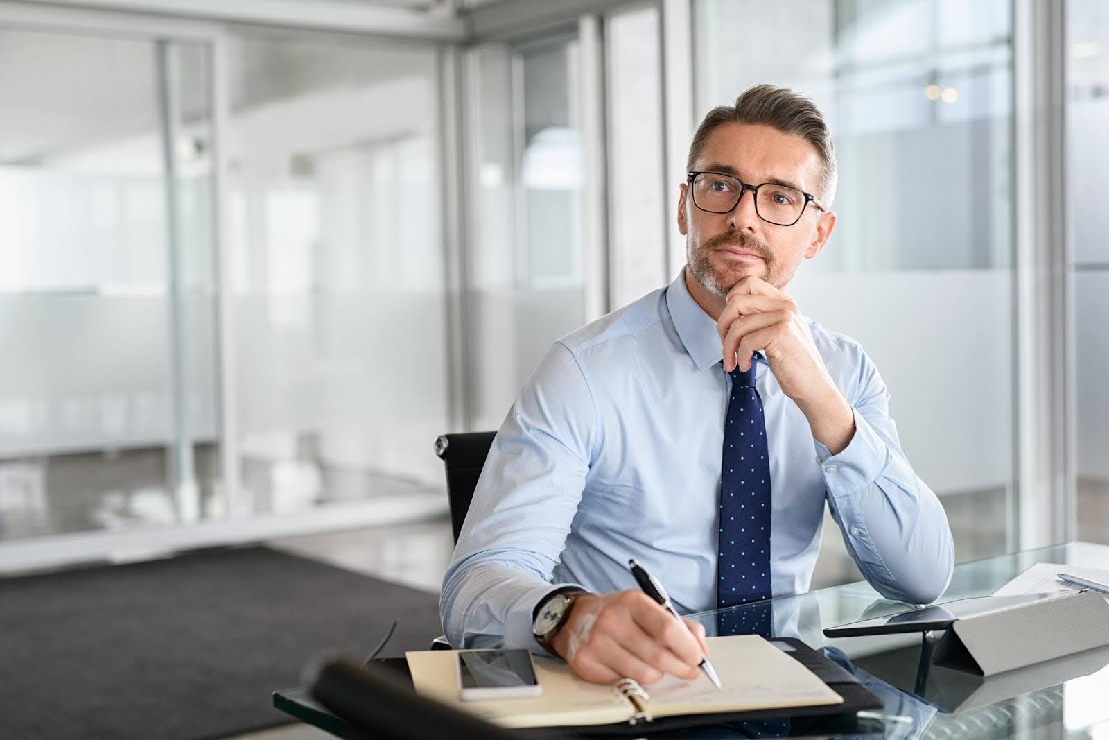 Confident businessman in an office setting writing in a notebook, reflecting on important business metrics and financial numbers to track weekly.