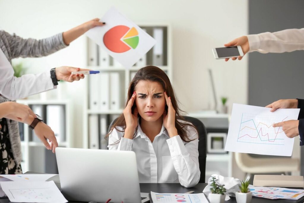 rustrated woman at her desk surrounded by coworkers handing her papers and phones, symbolizing feeling stuck in business due to taking on too many tasks alone.