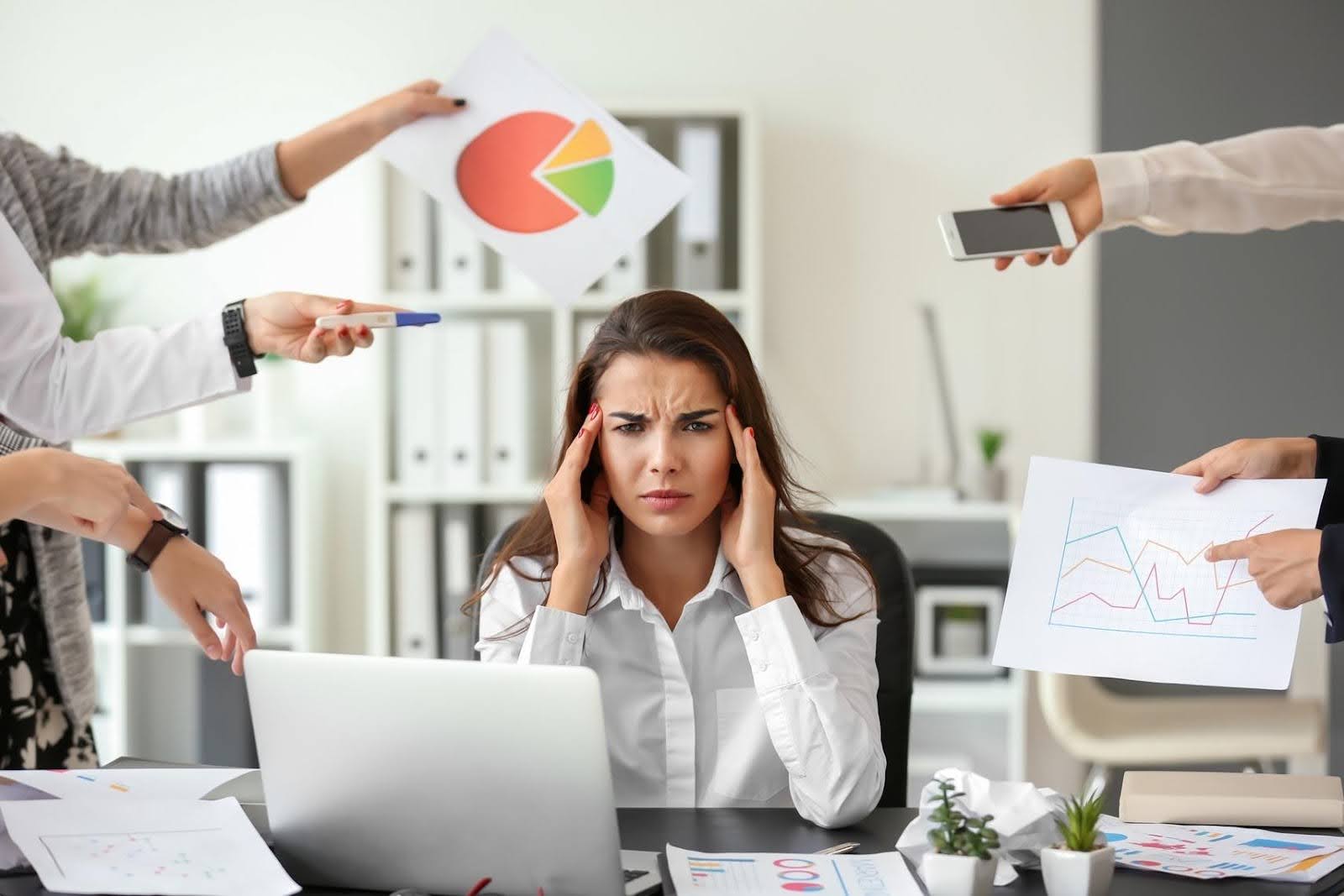 rustrated woman at her desk surrounded by coworkers handing her papers and phones, symbolizing feeling stuck in business due to taking on too many tasks alone.